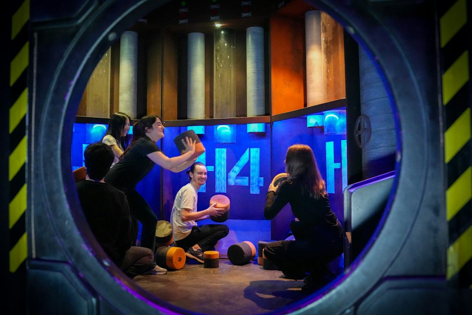 Quatre personnes manipulant des cylindres dans une salle d'action game futuriste, vus à travers un hublot.
