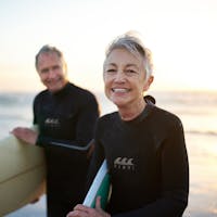 Middles aged husband and wife in wetsuits, holding surf boards on the beach.
