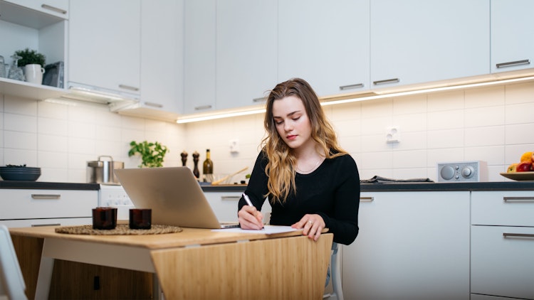 Nuori nainen tekee töitä kannettavalla tietokoneella vuokra-asunnon keittiön pöydän ääressä