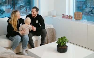 Matias Palmros ja Hanna-Riikka Nordman istuvat vaaleasävyisen kotinsa sohvalla ja hymyilevät toisilleen. Perheen pieni lapsi, Ava, on äidin sylissä. Taustalla on kaksi ikkunaa, joista näkyy lumisia mäntyjä.