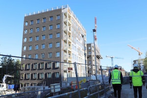 Linkkitorninkatu 7:n kaksi kerrostaloa rakentuvat Jaaninojan viereen.  Oikealla kävelee kaksi henkilöä, joilla  TVT:n ja Mangroven keltaiset turvaliivit.