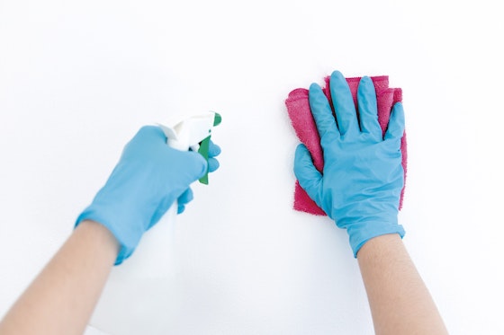 Kädet, joissa siniset kumihanskat, pesuainepullo ja siivousliina.