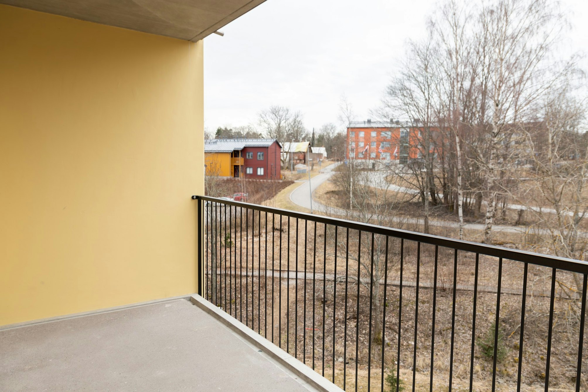 Parveke, jossa mustat metallikaiteet ja keltainen seinä. Parvekkeelta näkyy puita ja kauempana kerros- ja rivitaloja.
