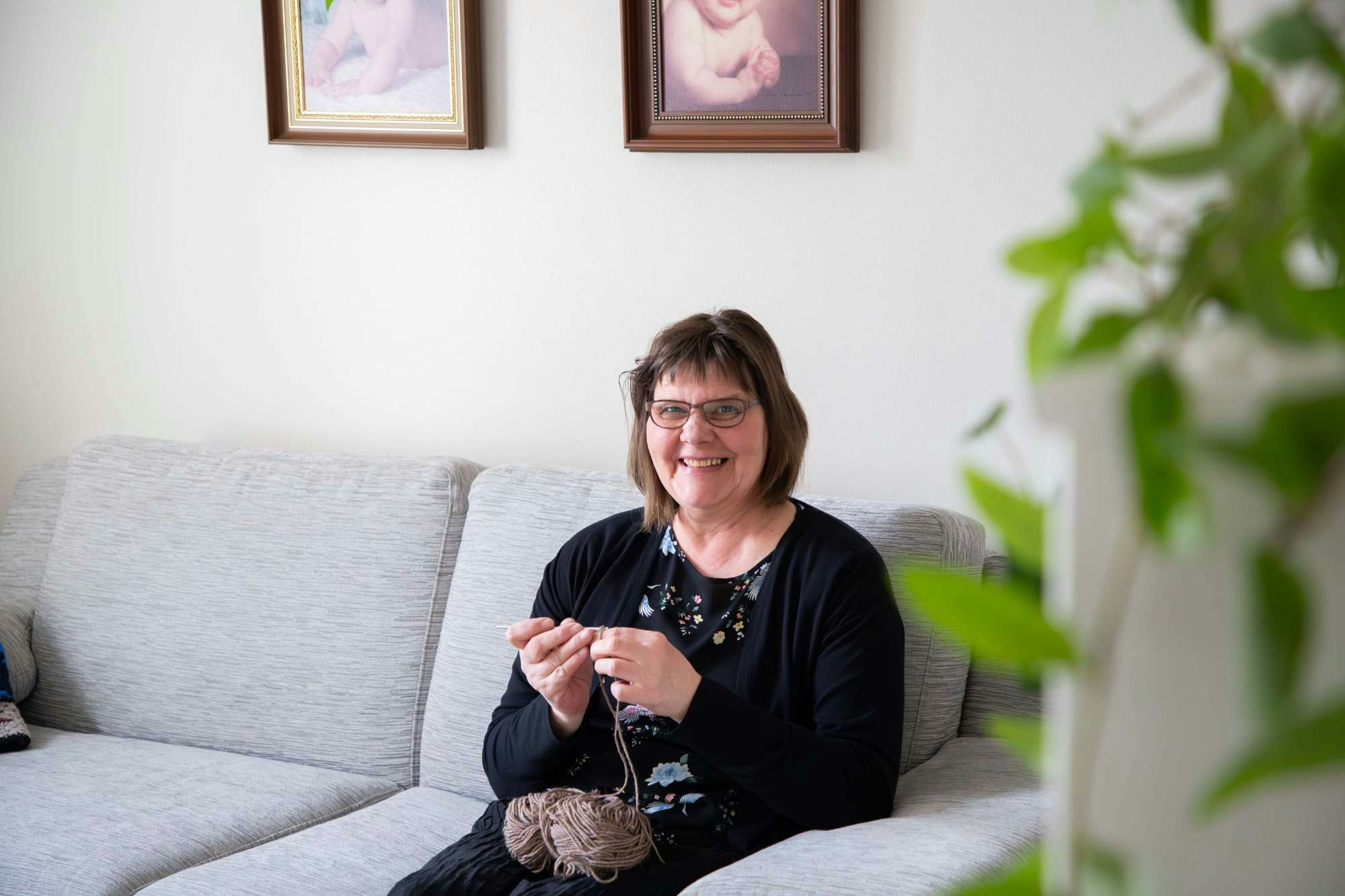 Anne-Maarit Reunanen istuu sohvalla kutomassa ja katsoo hymyillen kameraan.