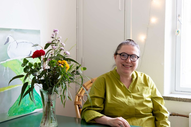 Kuvataiteilija Meri-Pauliina Sundén istuu keittiön pöydän ääressä ja hymyilee.