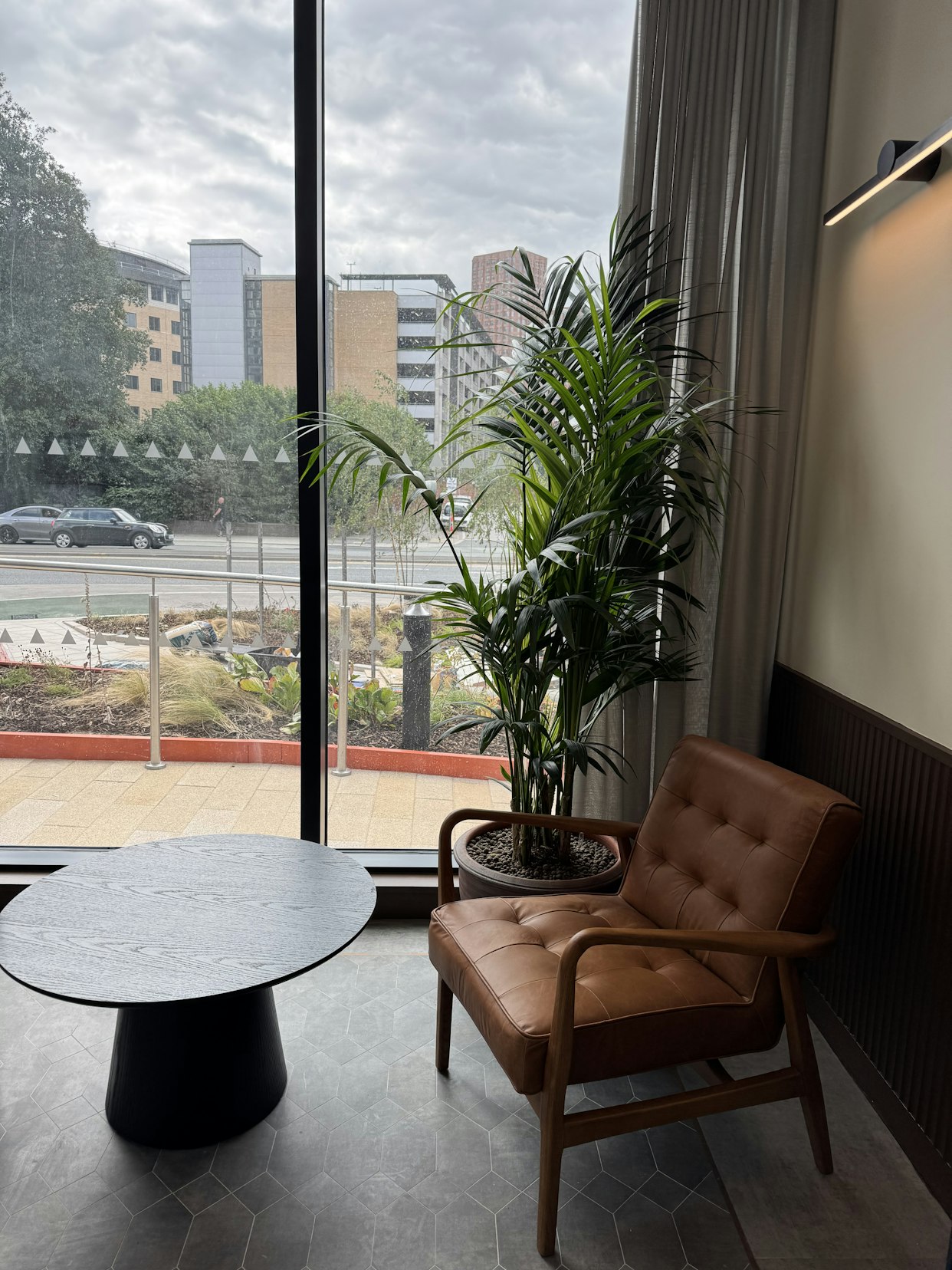 A tall potted palm plant next to a brown leather armchair and a round black side table by a large floor-to-ceiling window at Triangle Yard, Leeds