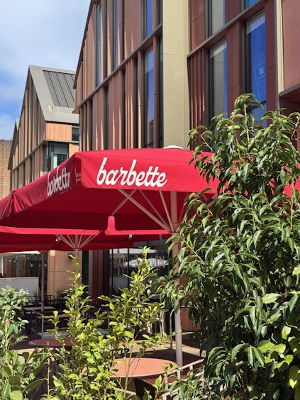 Lush green plants framing an outdoor dining area with bright red umbrellas bearing the Barbette logo, set against a modern brick building in Chester