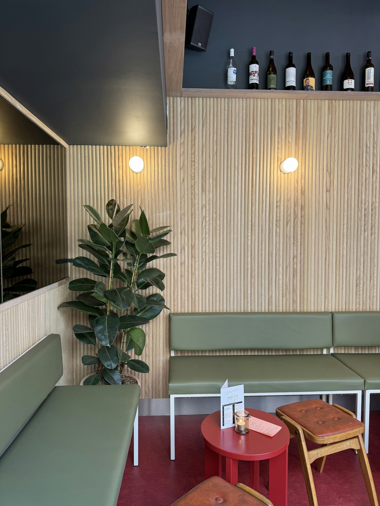 A leafy green potted plant situated next to green banquette seating against a light wood slatted wall, with small red cafe tables in the foreground at Barbette, Chester