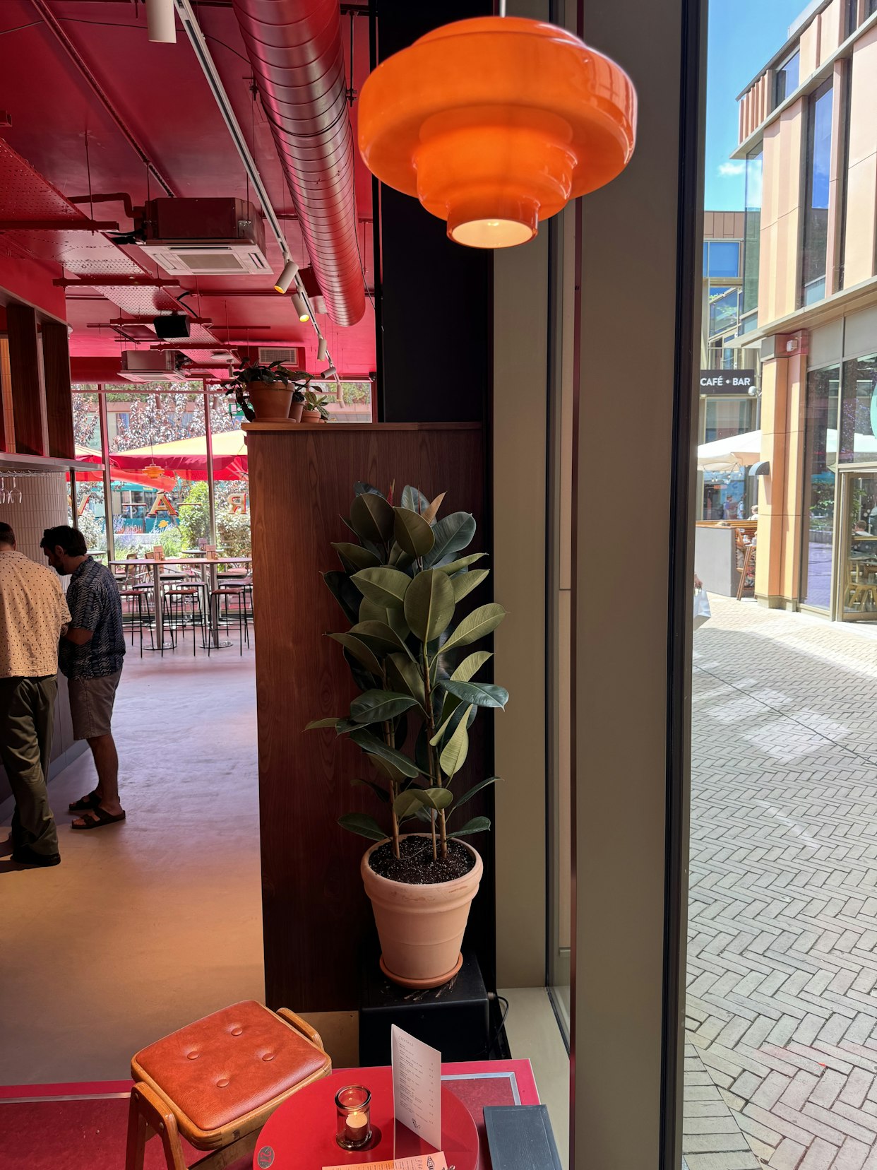 A potted green plant in a terracotta pot on a black stand, situated beneath a bright orange retro pendant lamp near a large window looking out onto the bustling outdoor terrace at Barbette, Chester