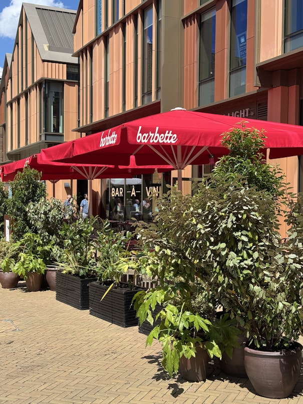A view of the Barbette outdoor dining area featuring bright red umbrellas, tables, and large black planters filled with lush green shrubs, set against a modern brick building facade.