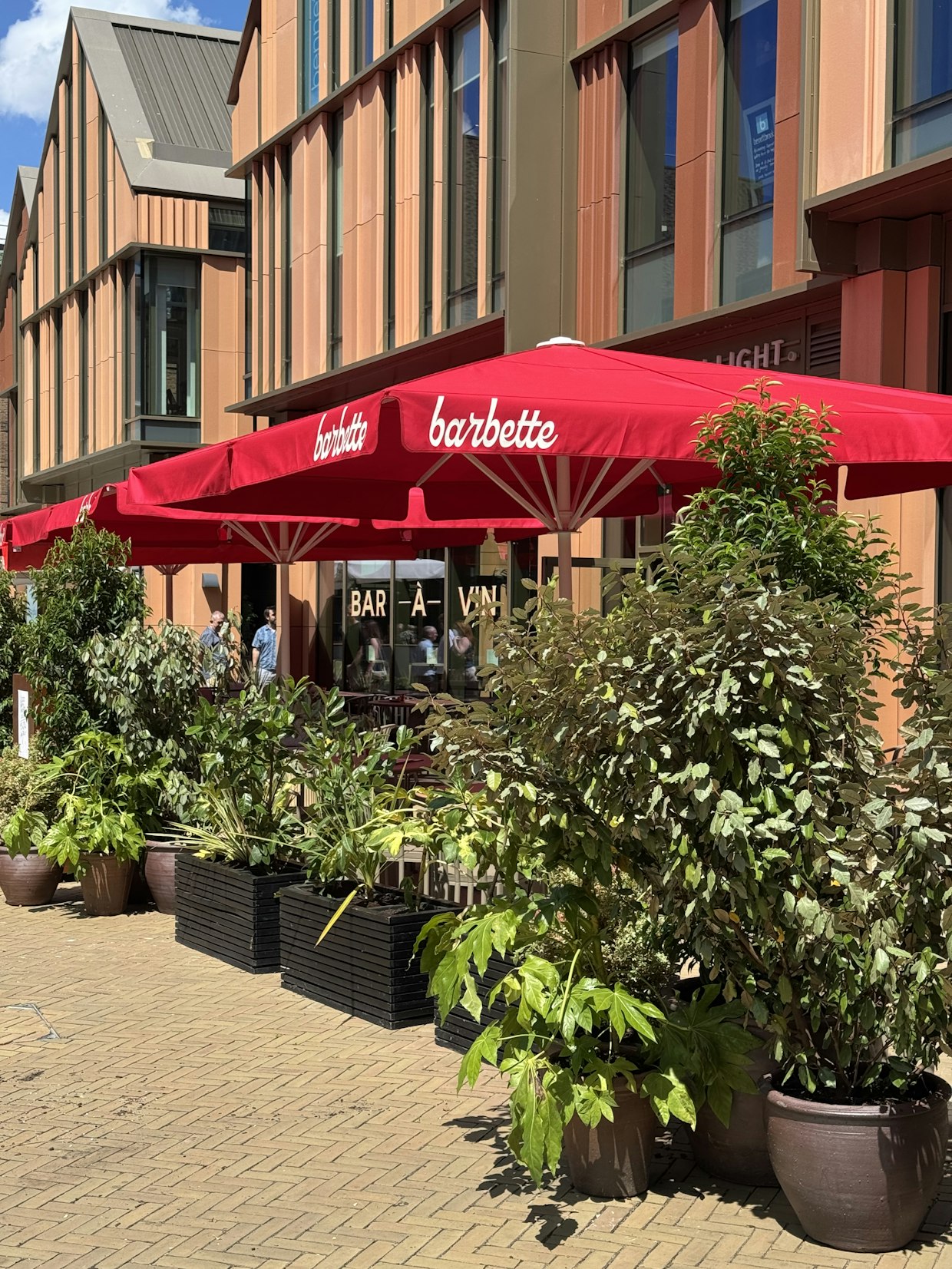 A view of the Barbette outdoor dining area featuring bright red umbrellas, tables, and large black planters filled with lush green shrubs, set against a modern brick building facade.