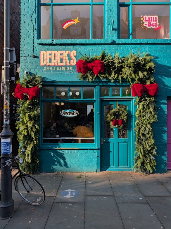 The vibrant teal exterior of Derek's Coffee & Sandwiches, decorated for Christmas with a large custom evergreen garland featuring red bows and ornaments framing the door and window.
