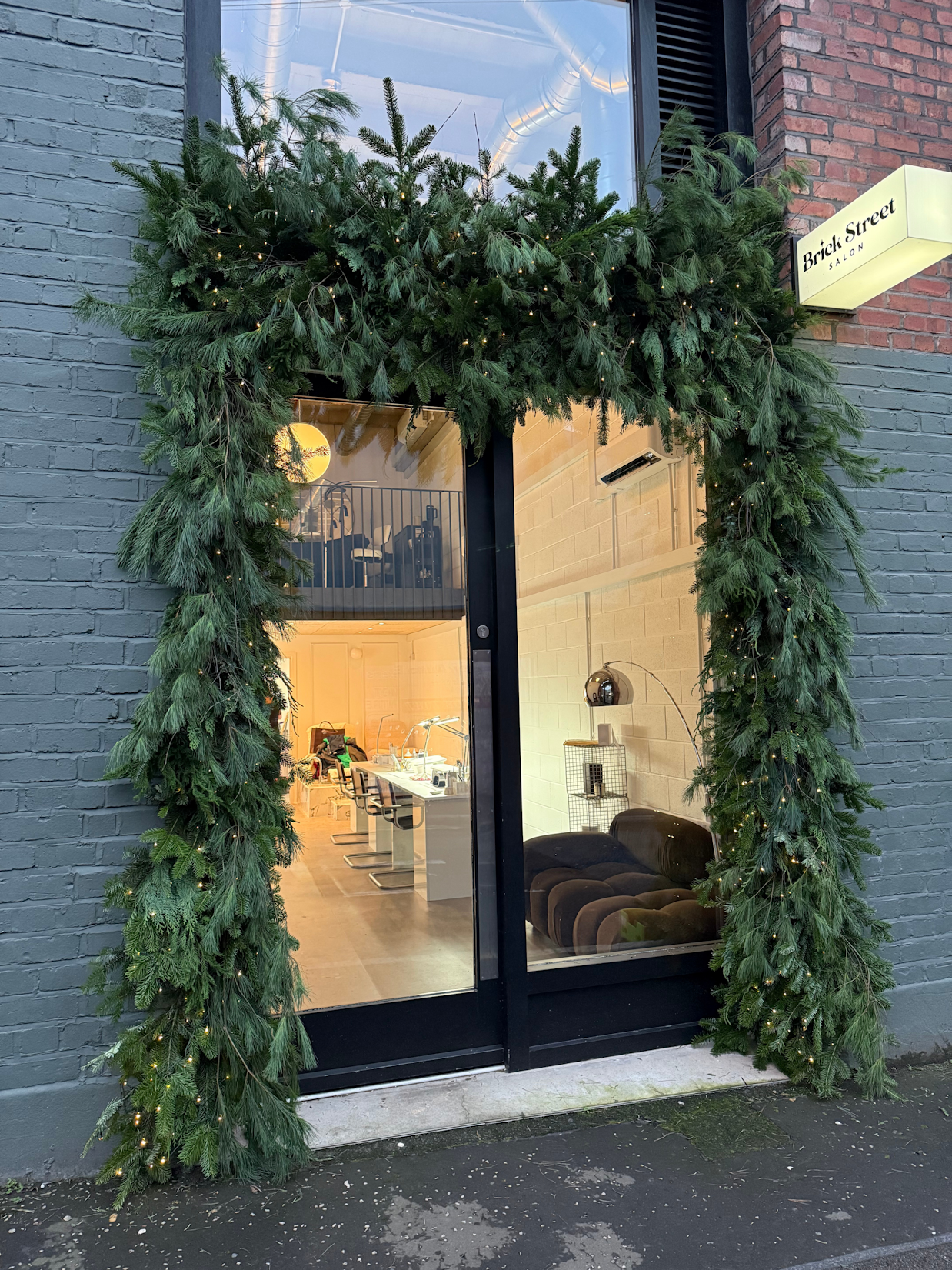 A lush green commercial Christmas garland woven with warm fairy lights, framing the glass entrance doors of the Brick Street salon against a dark brick exterior.