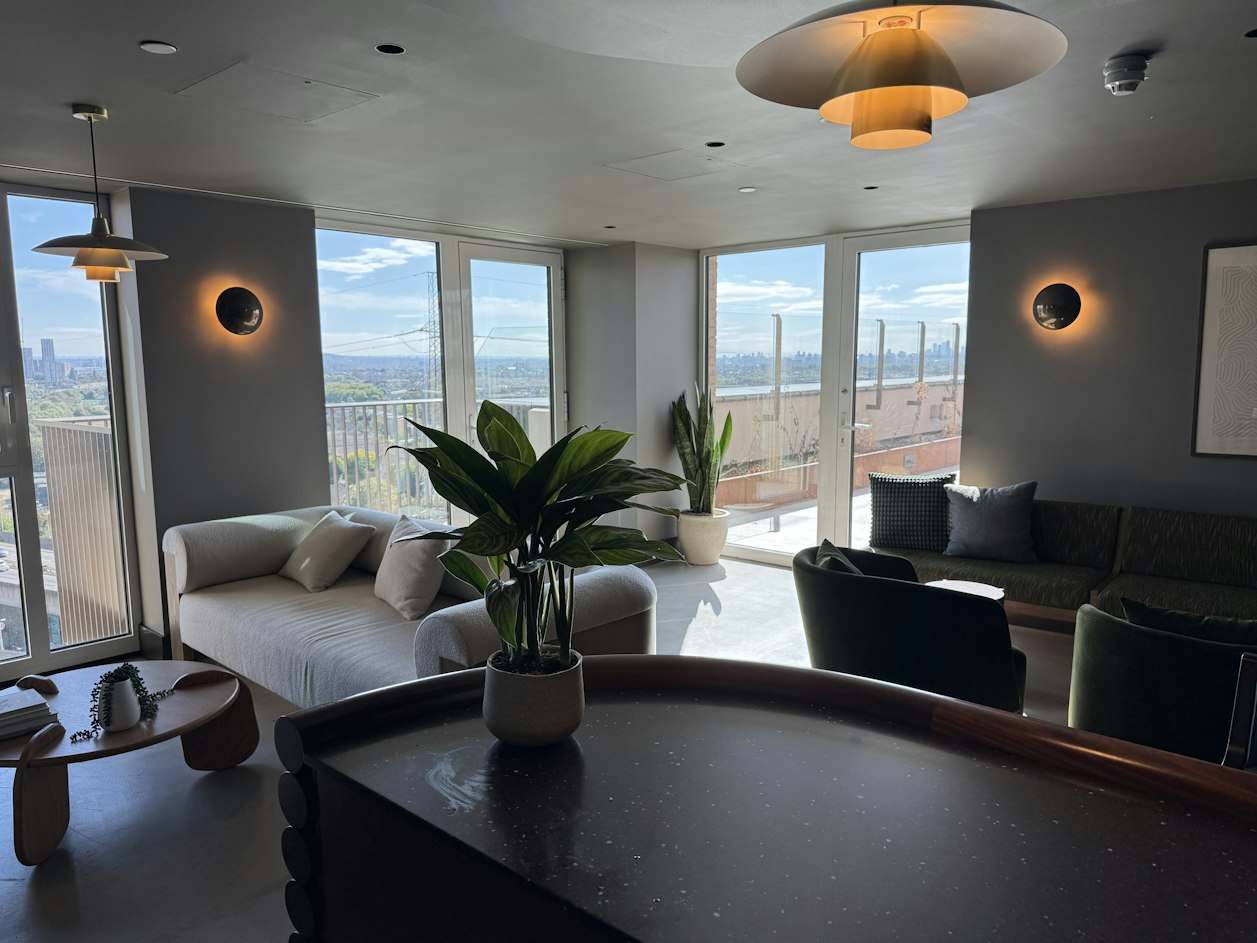 A modern lounge area in Aquifer House, London, featuring a potted plant on a dark curved table, comfortable seating, and large windows overlooking the city skyline.