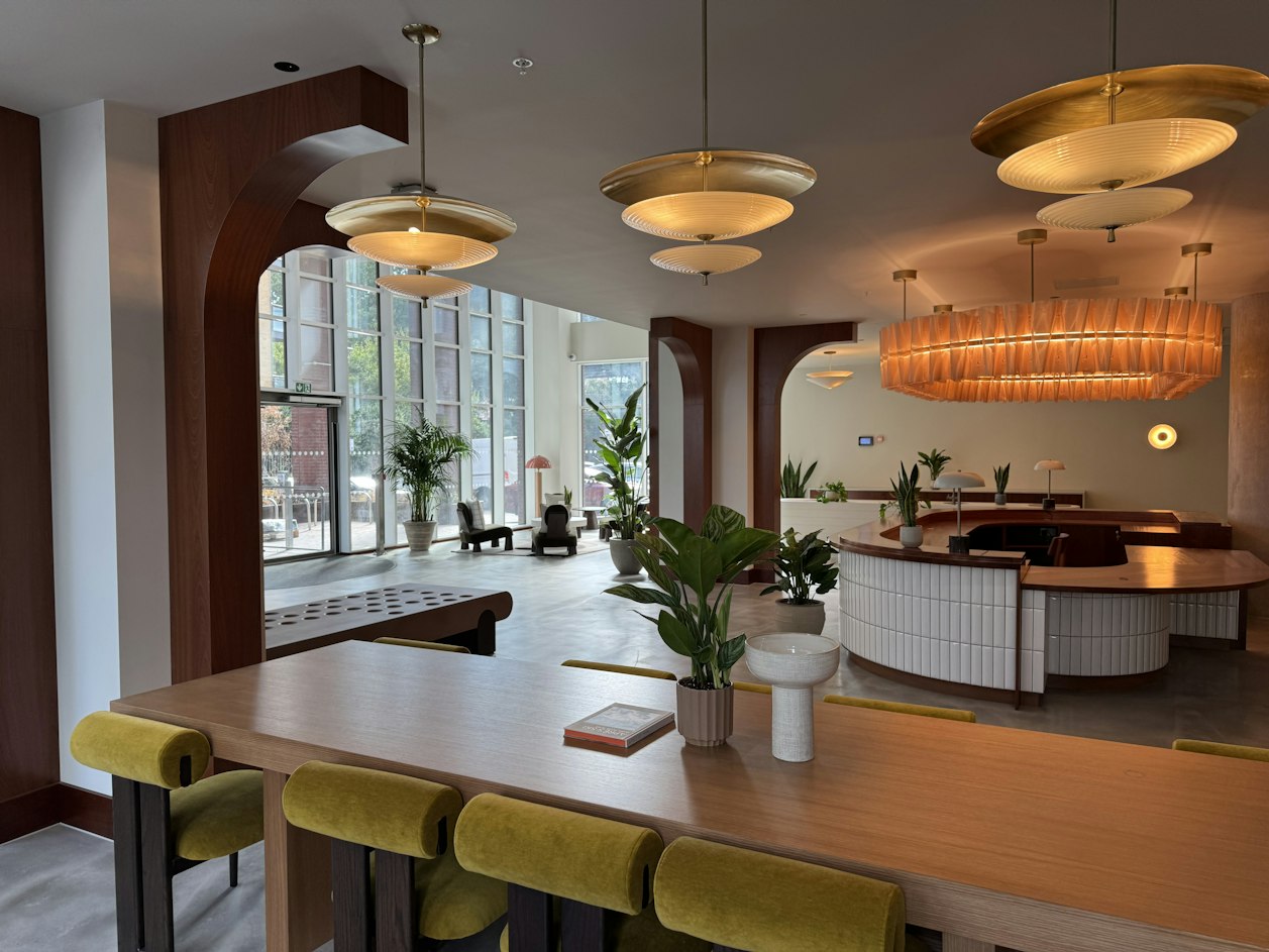 Potted greenery decorating a stylish London resident co-working and lounge area with a long wooden desk, yellow seating, and arched windows.