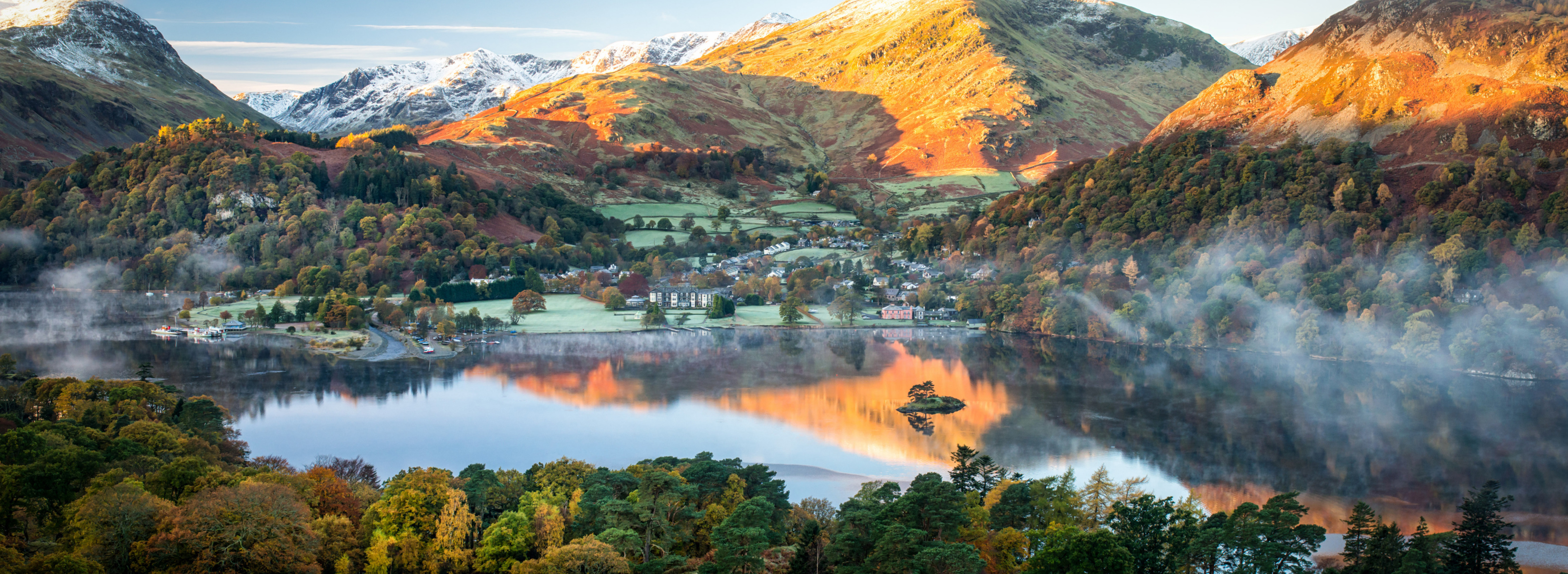Lake District with Misty lake surrounded by hills and snow-dusted mountains, with a small village on the shoreline