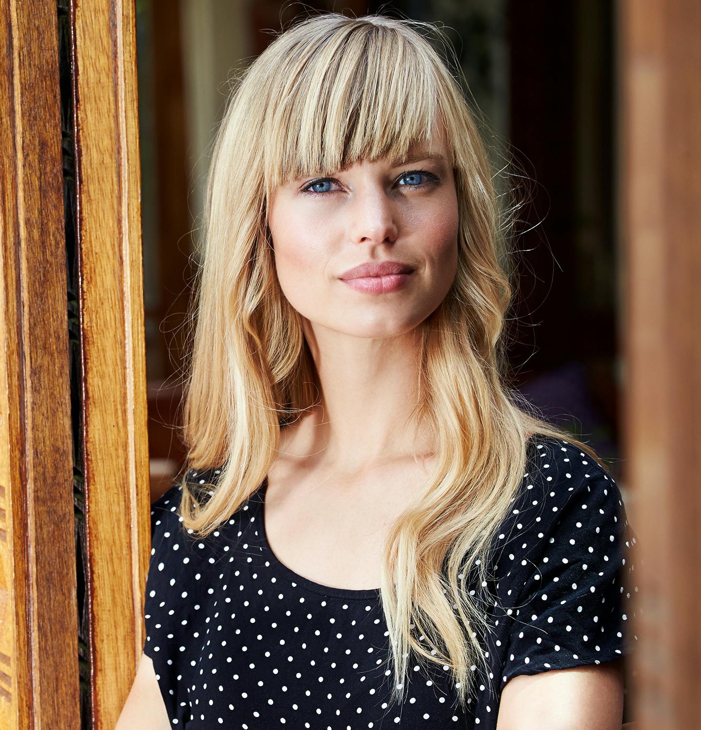 blond woman with blue eyes and bangs leaning against a wooden door