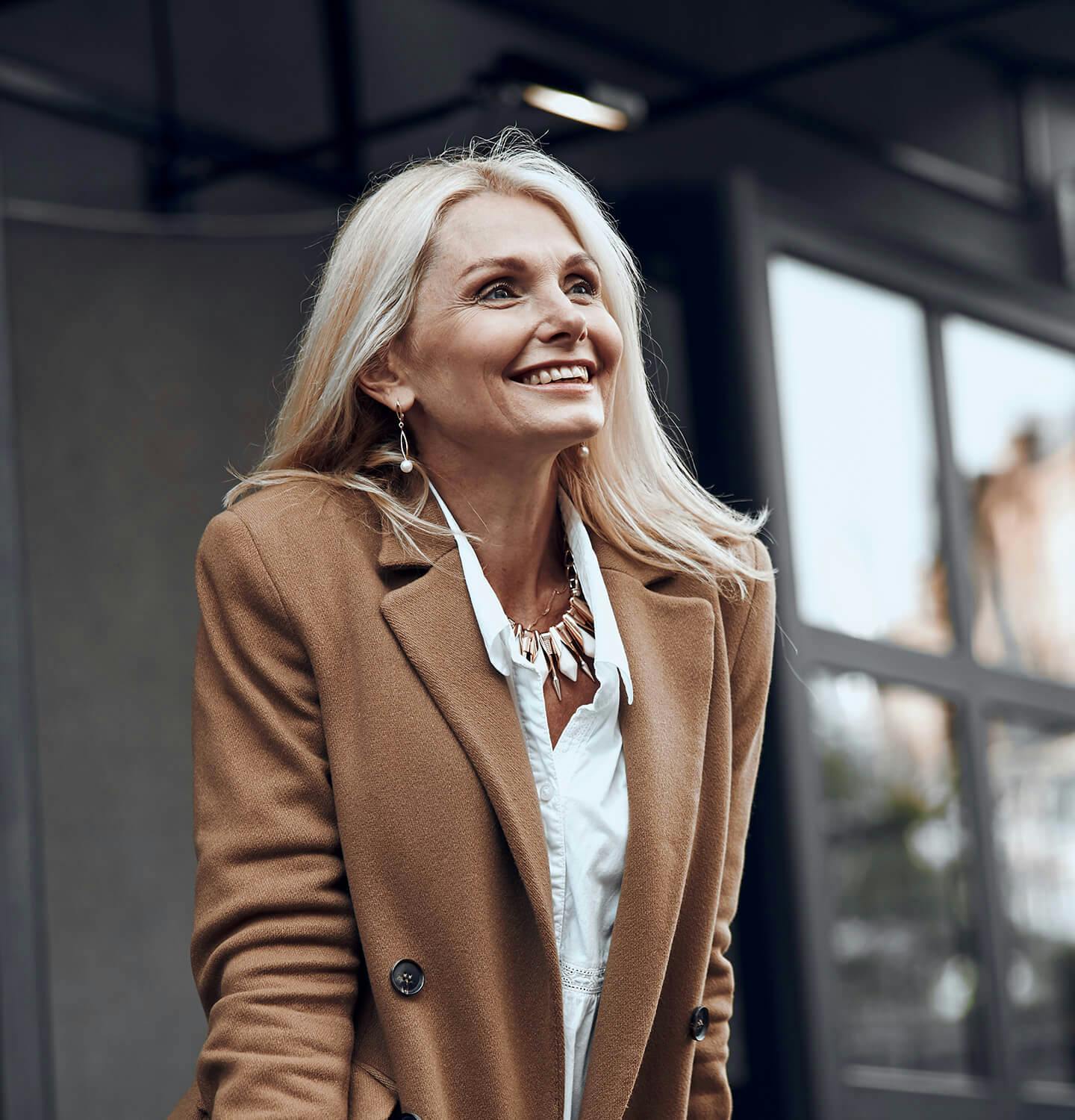 smiling woman in brown coat and white shirt standing outside of a building
