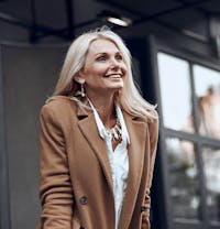 smiling woman in brown coat and white shirt standing outside of a building