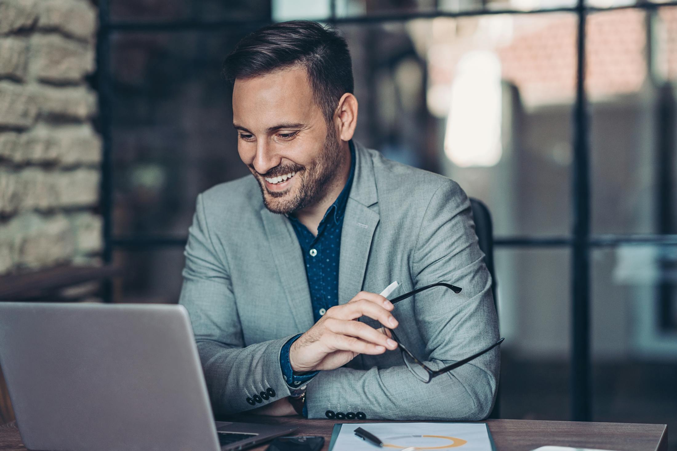 smiling man in grey suit sitting at a table with a laptop