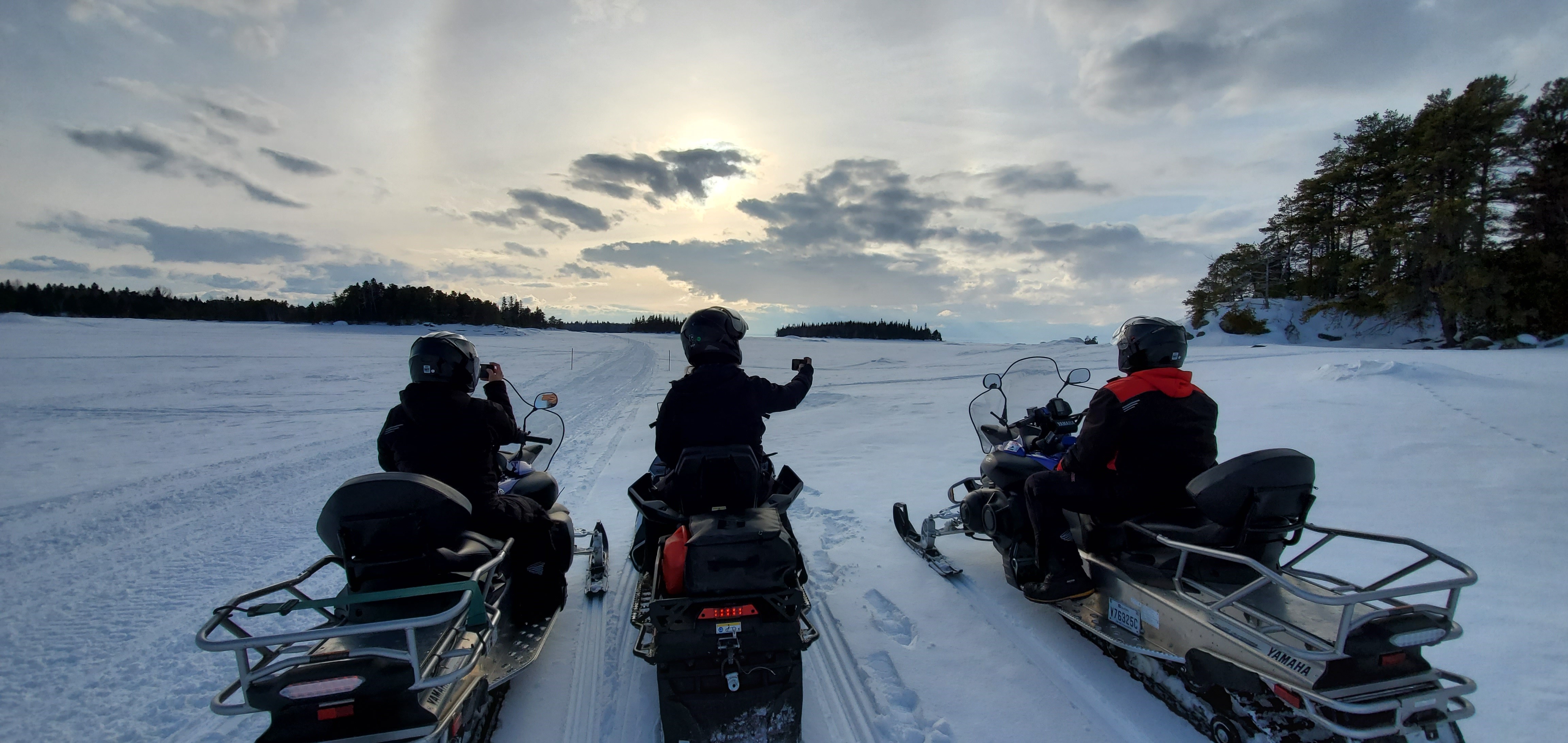 Trois motoneigustes sont arrêtés dans un décor enneigé pour photographier le nuage qui se cahe derrière les nuages, sur le lac Saint-Jean. À l'horizon, il y a les îles du parc national de la Pointe-Taillon.