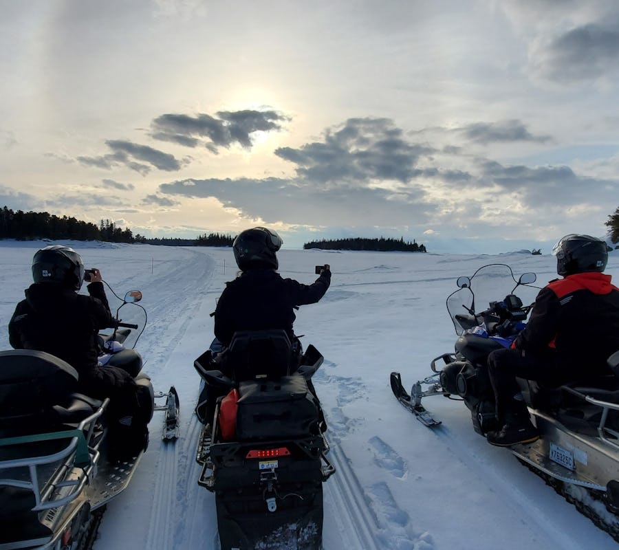 Trois motoneigustes sont arrêtés dans un décor enneigé pour photographier le nuage qui se cahe derrière les nuages, sur le lac Saint-Jean. À l'horizon, il y a les îles du parc national de la Pointe-Taillon.