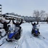 Un groupe de motoneigistes sont arrivés au centre Équinox Aventure à l'Auberge des îles et manifestent leur joie en levant les bras dans les airs. Derrière on voit l'Auberge des îles enneigée.