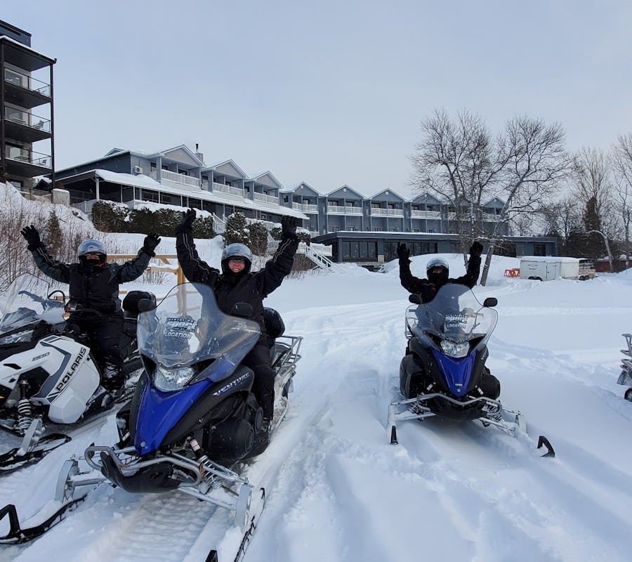 Un groupe de motoneigistes sont arrivés au centre Équinox Aventure à l'Auberge des îles et manifestent leur joie en levant les bras dans les airs. Derrière on voit l'Auberge des îles enneigée.