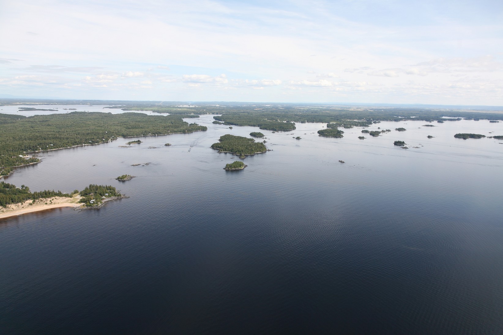 La vue érienne du lac Saint-Jean est impressionante ! L'eau semble calme, des îles peuplées d'arbres et de plages sabloneuses alimentent le paysage à perte de vue. Le ciel est clair avec quelques nuages blancs. La nature et la sensation de grandeur du lac Saint-Jean est saisissante. The aerial view of Lake Saint-Jean is impressive! The water seems calm, islands populated with trees and sandy beaches fill the landscape as far as the eye can see. The sky is clear with a few white clouds. The nature and feeling of grandeur of Lake Saint-Jean is striking.