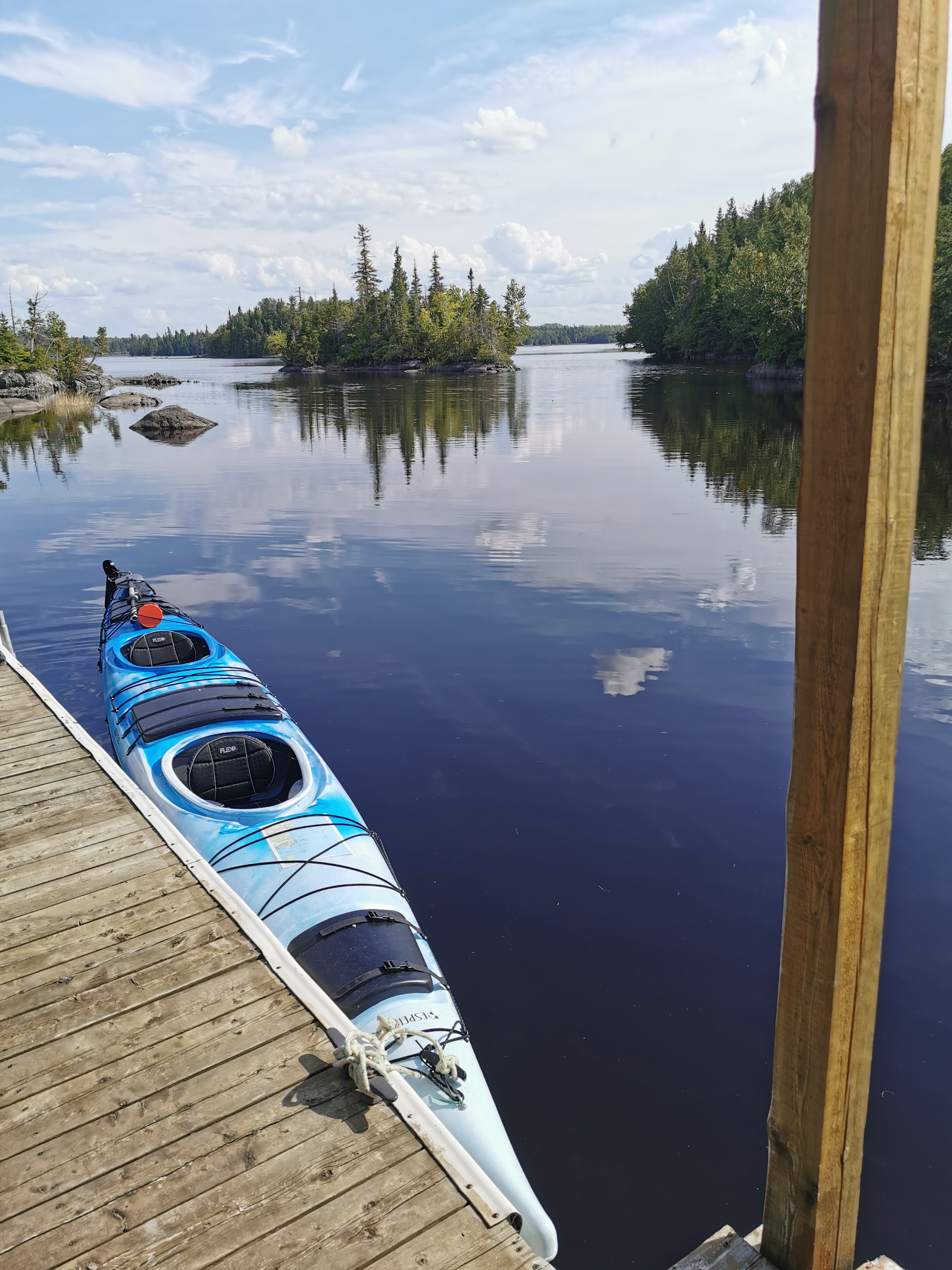 Voici la vue de la terrasse flottante de l'iloft. L'eau est calme, il fait beau et on voit des petites îles avec des grands feuillus verts. Un kayak de mer duo est accosté à la terrasse flottante. Here is the view from the floating terrace of the iloft. The water is calm, the weather is nice and we can see small islands with large green deciduous trees. A duo sea kayak is docked at the floating terrace.