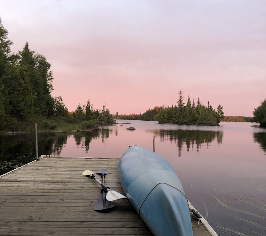 Voici la vue qu'on a sur la terrasse flottante de l'iloft au coucher du soleil. Le ciel est rose et se reflête sur le lac miroir. On voit les petites îles autour avec leurs grands feuillus verts. Un kayak est retourné sur la terrasse pour éviter que les bancs se mouiller par la rosée. Here is the view we have on the floating terrace of the iloft at sunset. The sky is pink and reflected on the mirror lake. We see the small islands around with their large green deciduous trees. A kayak is returned to the terrace to prevent the benches from getting wet with dew.