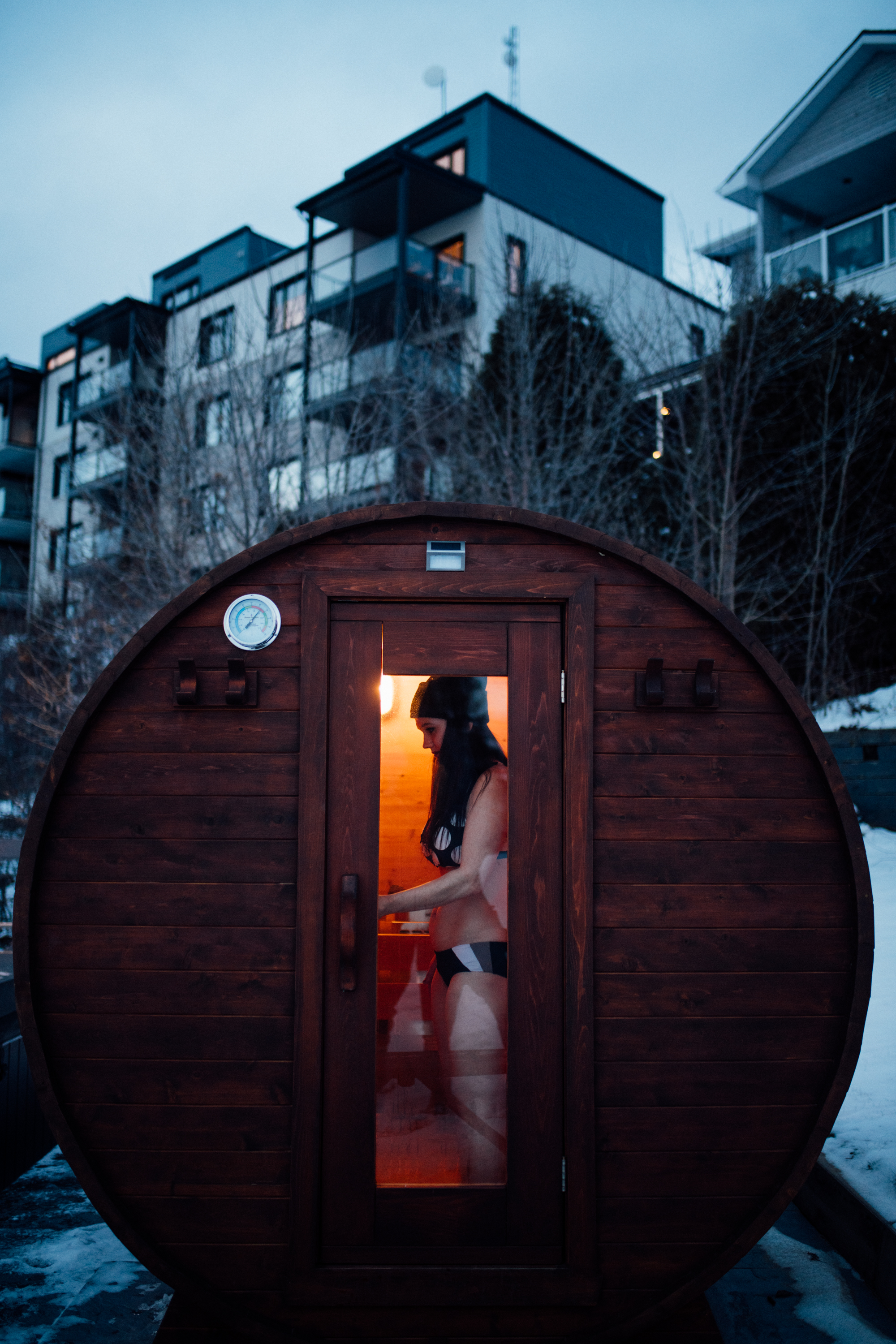 Au premier plan, un sauna extérieur rond en bois foncé, avec une personne debout à l'intérieur. La lumière à l'intérieur est chaleureuse. À l'arrière-plan, vue sur l'auberge des îles avec des balcons visibles.