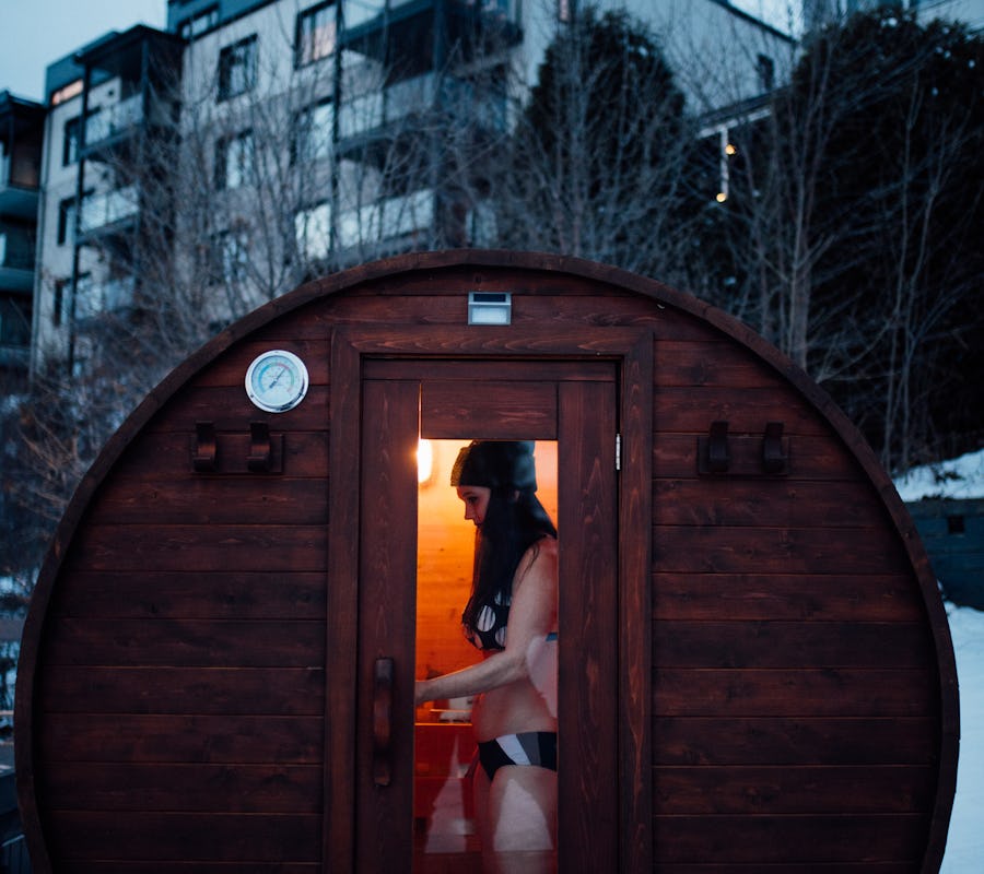 Au premier plan, un sauna extérieur rond en bois foncé, avec une personne debout à l'intérieur. La lumière à l'intérieur est chaleureuse. À l'arrière-plan, vue sur l'auberge des îles avec des balcons visibles.