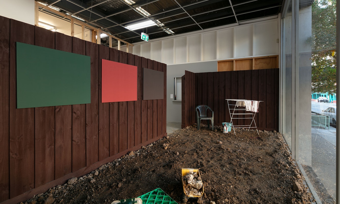 Clinton Naina, Heritage Colours, 2000/2025, installation view, 1964, 1969, 1977, 1995, 2000, 2005, 2025, Gertrude Contemporary, Naarm Melbourne, 2025, soil, drop sheet, painted timber fence, laser prints, plastic chair, heater, clothes horse, milk crate, paint, projection, image courtesy the artist and Gallerysmith, Naarm Melbourne © the artist, photograph: Christian Capurro.
