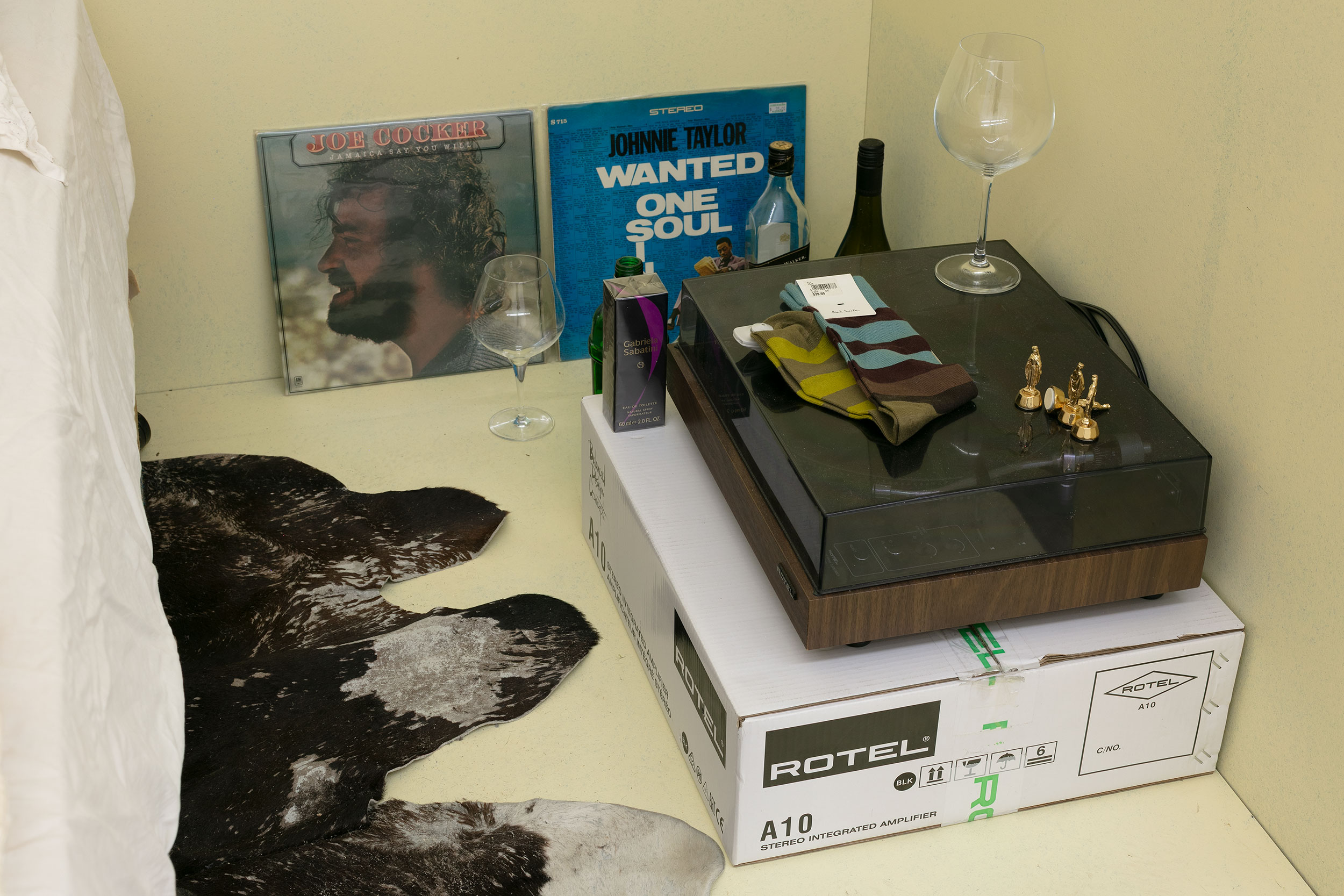 Lane Cormick, Vanilla Sigartje (detail), 2002/2025, installation view, 1964, 1969, 1977, 1995, 2000, 2005, 2025, Gertrude Contemporary, Naarm Melbourne, 2025, cow hide, bed, doona, pillow, MDF, wall paint, suit, coat hanger, ink on paper, record sleeves, glass bottles, glasses, Gabriela Sabatini  eau de toilette, Paul Smith socks, cardboard box, metal Virgin Marys, image courtesy the artist and Daine Singer, Naarm Melbourne© the artist, photograph: Christian Capurro.