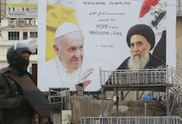 An Iraqi security guard stands in front of a billboard bearing portraits of the Pope and Ayatollah Ali Sistani in central Baghdad on March 4, 2021. (Photo via Getty Images)