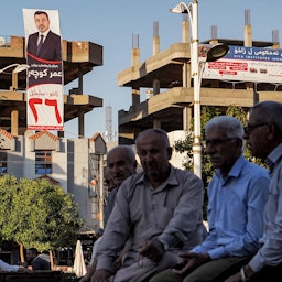 چند مرد، نشسته در نزدیکی ساختمانی که بنر تبلیغاتی انتخابات پارلمانی ۲۸ مهر/ ۱۰ اکتبر بر روی آن نصب شده است؛ زاخو، ۱۳ مهر ۱۴۰۰/ ۵ اکتبر ۲۰۲۱. (عکس از گتی ایمیجز)