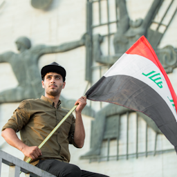 پرچم عراق در دستان یک معترض؛ میدان تحریر، بغداد، ۵ مرداد ۱۳۹۷/ ۲۷ ژوئیه ۲۰۱۸. (عکس از مصطفی نادر/ ویکی‌مدیا کامنز)
