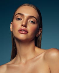 Woman with blonde hair is having portrait taken with blue background. Her face is glowing and looks healthy to show the possible results with using ZO Skincare