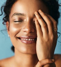 Woman having portrait taken with blue background. One hand is almost cover her left side of ehr face and she is smiling with her eyes closed.Her face is glowing and looks healthy to show the possible results with using ZO Skincare