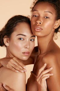 Two women in their 20's are having her portrait taken together. Their skin is glowing as they looks directly at the camera. Their glowing skin is to emulate the potential results of using the Acorn Treatment