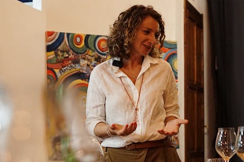 Claudia Piras parla e gesticola durante una conversazione, con alcuni calici di vino in primo piano.