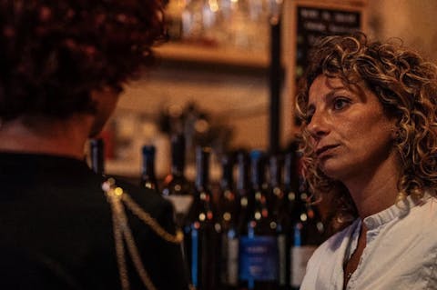 Claudia Piras conversa con un’altra persona in un ambiente di ristorazione, tra bottiglie di vino.