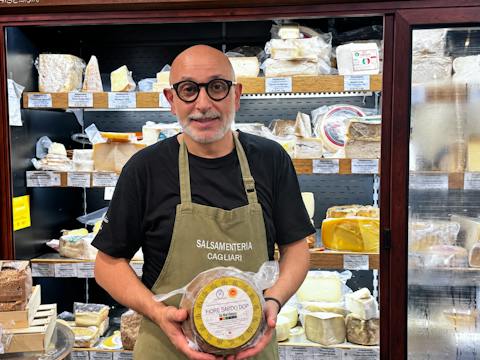 Michele Cherchi in a cheese shop in Cagliari, standing beside a counter displaying a selection of Sardinian cheeses