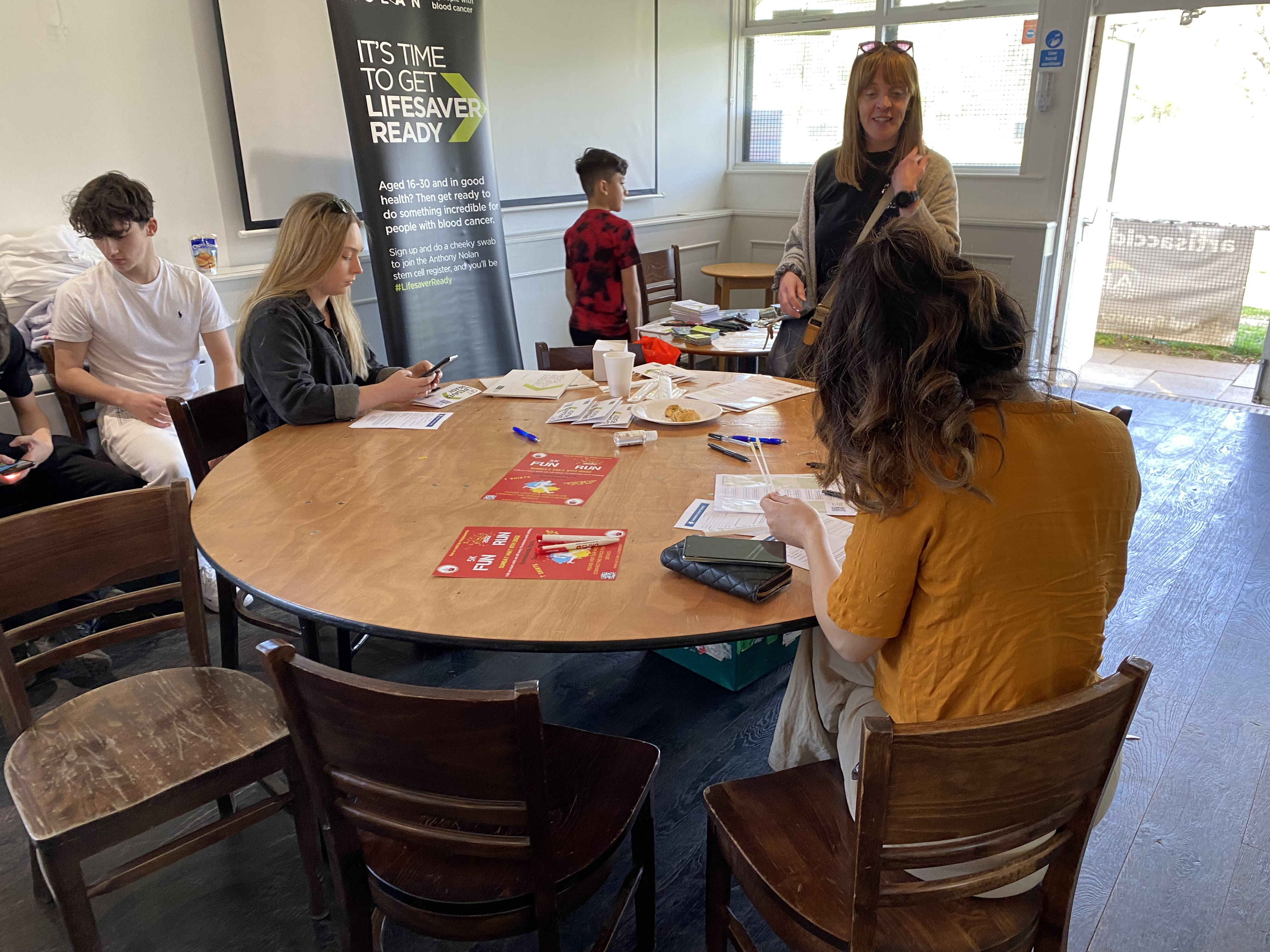 Young people fill in forms at a round table, Anthony Nolan banner in the background