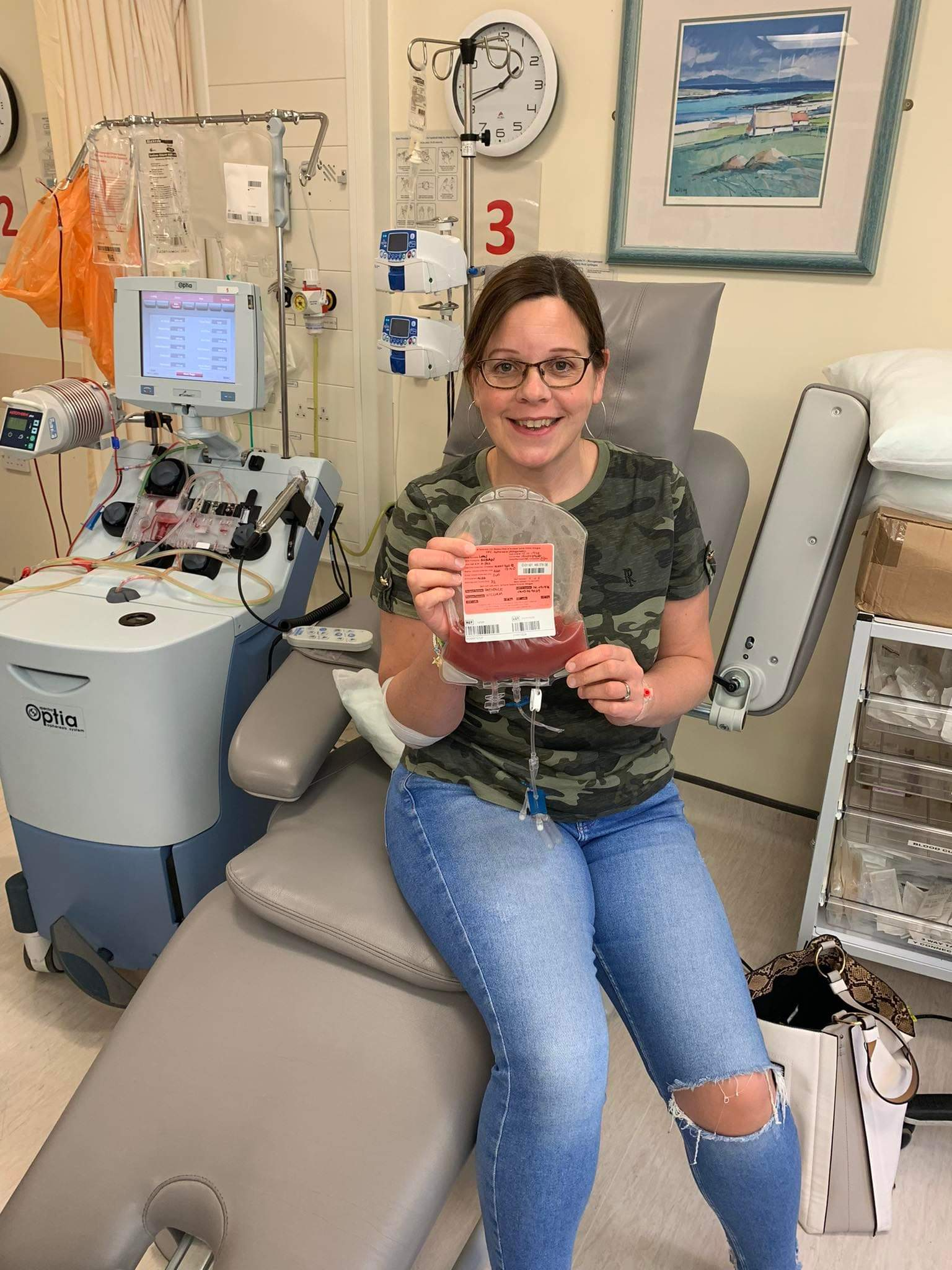 White woman sat on the edge of a hospital bed in jeans and a black top  holding a blood bag. 