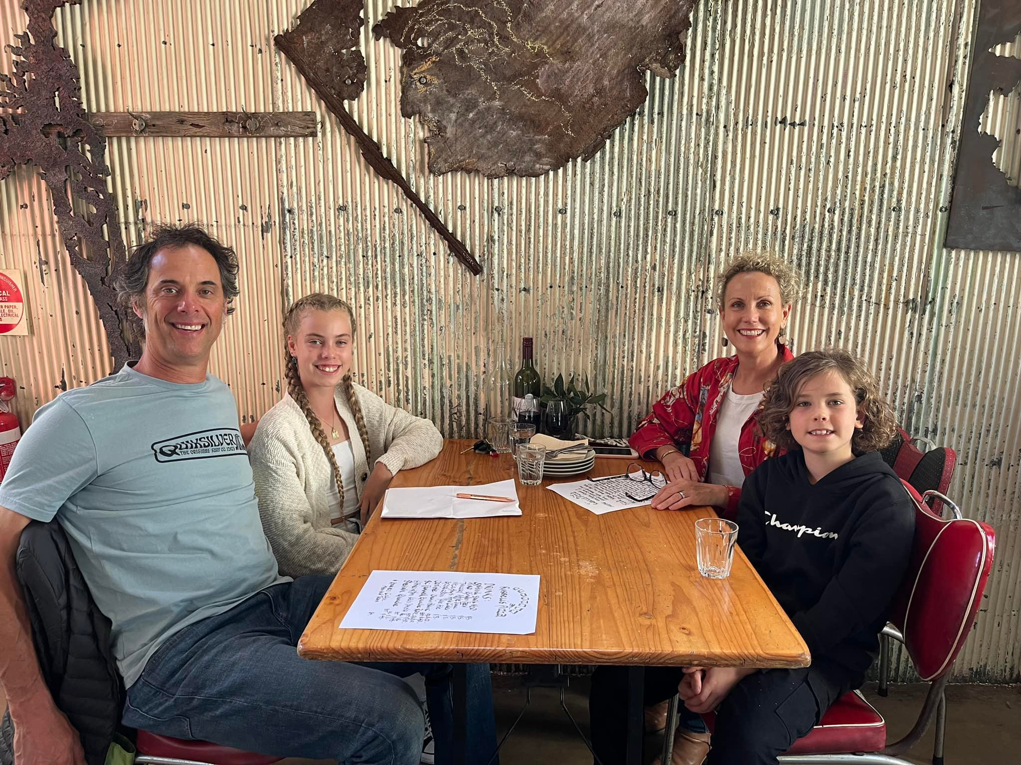 A family of four sat at a cafe table all smiling at the camera. At the back, Tania is sat across from her daughter. Her son  sits next to her and opposite him is her husband. They all look relaxed and happy.