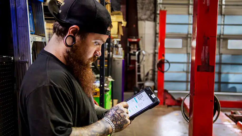Man with tattoos, gauges and red beard holding a tablet using Shopmonkey while standing in an auto shop.