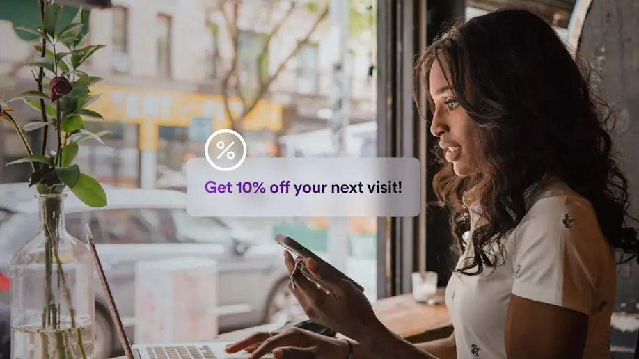 A woman on a laptop getting a notification on her phone with a discount offer.