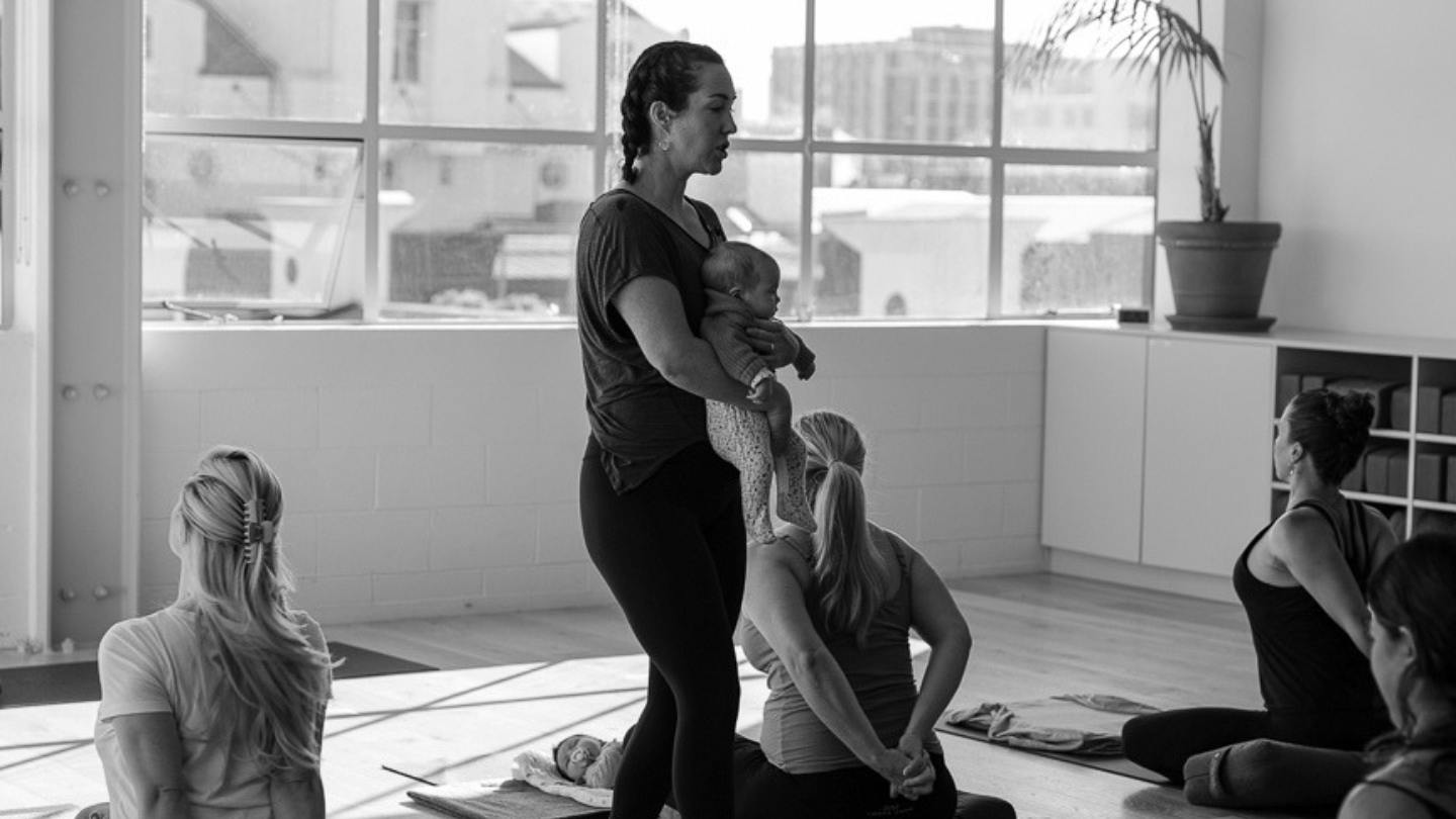 Julia Wilkinson holding a baby on her hip while leading a group class of women on exercise mats